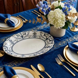 K&ouml;nnte beinhalten: Eine formelle Tischdekoration mit einer blauen Samt-Tischdecke, goldger&auml;nderten Tellern und goldenem Besteck. Eine gro&szlig;e ovale Platte in Wei&szlig; und Blau steht im Mittelpunkt. Blaue Servietten werden von goldenen Serviettenringen gehalten. Blumen und Kerzen.