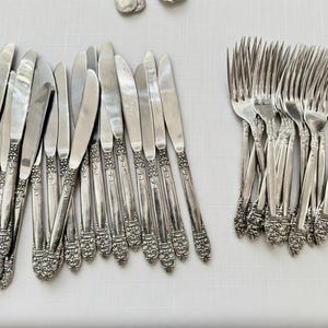 May include: A collection of vintage silverware, including knives and forks, arranged on a white surface. The silverware has ornate, detailed handles and a shiny, reflective finish, suggesting a formal dining set.