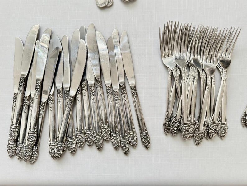 May include: A collection of vintage silverware, including knives and forks, arranged on a white surface. The silverware has ornate, detailed handles and a shiny, reflective finish, suggesting a formal dining set.