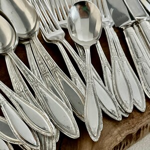 May include: A set of silver flatware, including spoons, forks, and knives, with ornate designs. The flatware is arranged in a fan shape on a wooden surface.