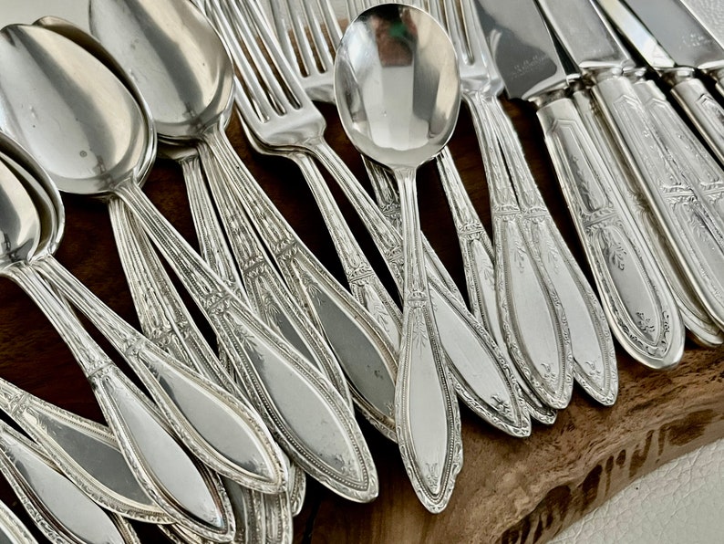 May include: A set of silver flatware, including spoons, forks, and knives, with ornate designs. The flatware is arranged in a fan shape on a wooden surface.