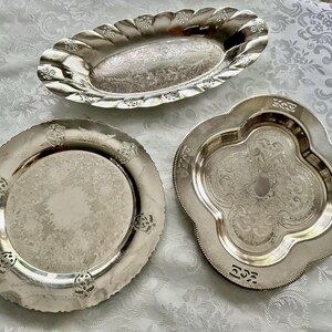 May include: Three silver serving trays with ornate designs. The trays are oval, round, and quatrefoil-shaped. The oval tray has a scalloped edge. The round tray has a decorative edge with floral accents. The quatrefoil tray has a floral design.