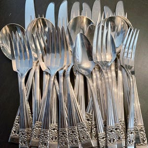 May include: A collection of silver-toned stainless steel flatware including forks, spoons, and knives. Each piece features a polished finish with a vertical ribbed pattern on the handle and a detailed floral embossed design at the base.