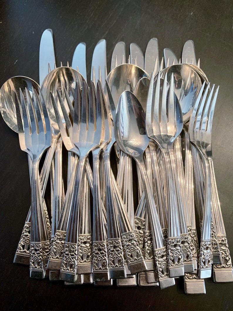 May include: A collection of silver-toned stainless steel flatware including forks, spoons, and knives. Each piece features a polished finish with a vertical ribbed pattern on the handle and a detailed floral embossed design at the base.
