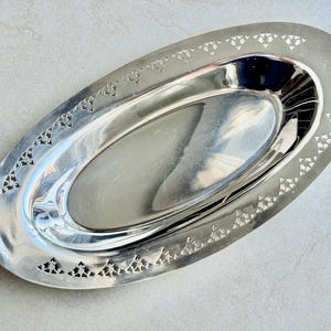 K&ouml;nnte beinhalten: Ein ovales Serviertablett aus silberfarbenem Metall mit einer polierten, reflektierenden Vertiefung in der Mitte. Der breite, flache Rand weist ein sich wiederholendes, ausgestanztes Muster aus kleinen Dreiecken auf.