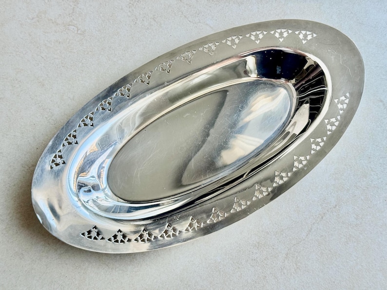K&ouml;nnte beinhalten: Ein ovales Serviertablett aus silberfarbenem Metall mit einer polierten, reflektierenden Vertiefung in der Mitte. Der breite, flache Rand weist ein sich wiederholendes, ausgestanztes Muster aus kleinen Dreiecken auf.