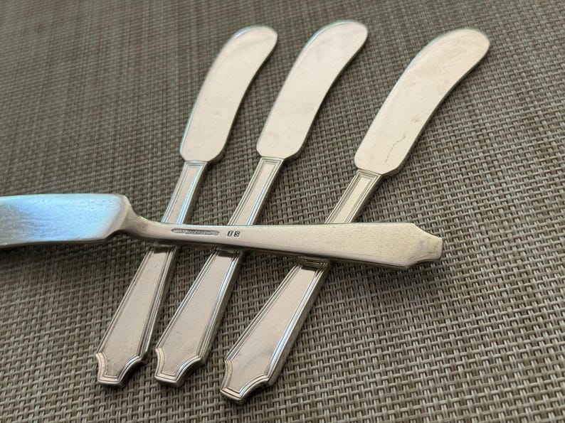 May include: A set of silver butter knives, each with a curved blade and a decorative handle. The knives are arranged on a textured surface, with one knife overlapping the others. The image focuses on the silverware's design and material.