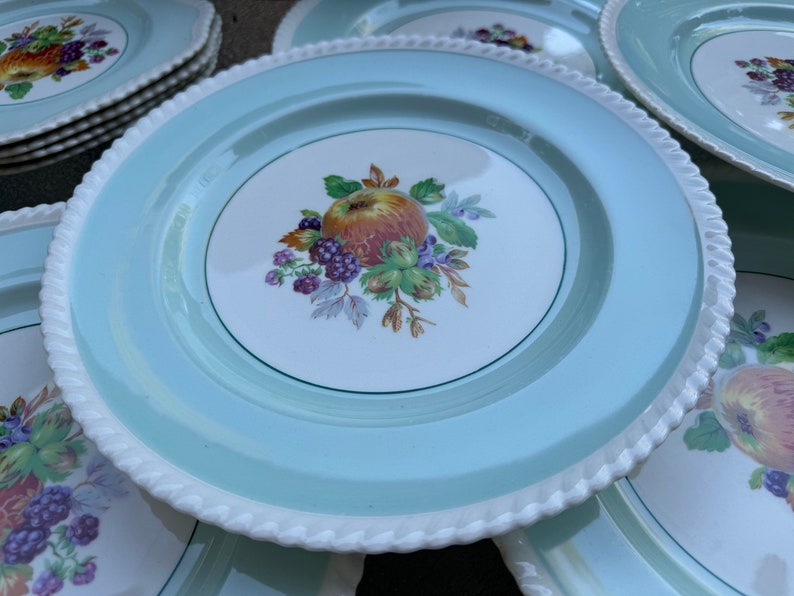 Puede incluir: Una pila de platos de cena de cer&aacute;mica azul claro con un borde blanco en forma de cuerda. Cada plato presenta un centro blanco con un borde verde y un dise&ntilde;o pintado de una manzana roja, moras y hojas verdes.