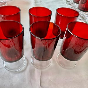 May include: Set of eight red glass wine glasses with clear stems and bases. The glasses are arranged on a white tablecloth.