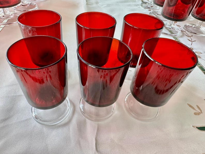 May include: Set of eight red glass wine glasses with clear stems and bases. The glasses are arranged on a white tablecloth.