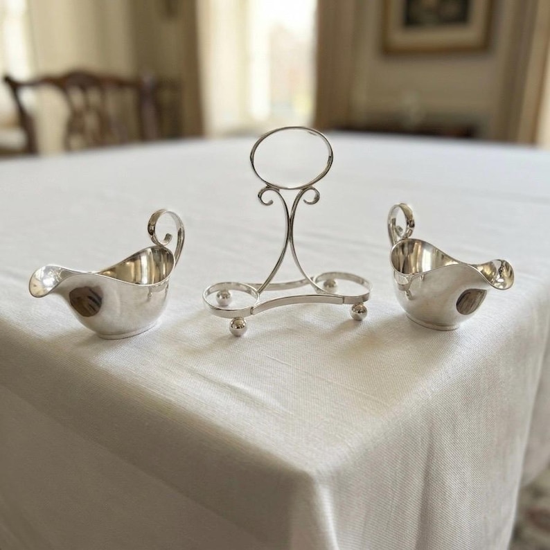 Puede incluir: Un conjunto de tres objetos de metal plateado que consta de dos peque&ntilde;os recipientes vertedores curvos con asas y un soporte central con un aro superior circular, brazos de soporte curvos y tres pies esf&eacute;ricos sobre una base triangular.