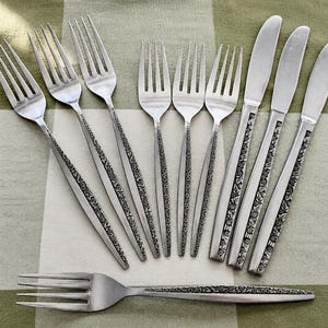 May include: A set of stainless steel flatware consisting of six forks and three knives arranged in a row, with one additional fork placed horizontally below. Each piece features a detailed floral pattern embossed along the handle.