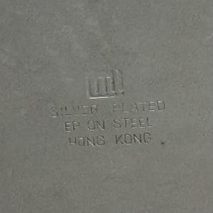 Puede incluir: Una superficie de metal de color plateado con un logotipo en relieve y el texto: SILVER PLATED, EP ON STEEL, HONG KONG. Las letras est&aacute;n estampadas directamente en el material met&aacute;lico, mostrando una marca de origen clara.