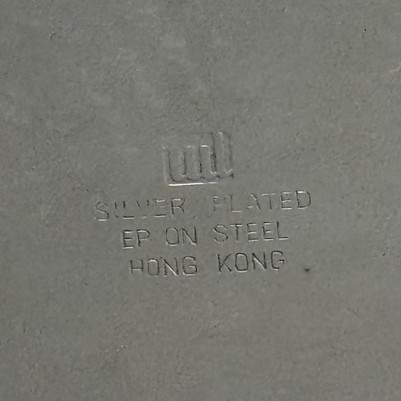 Puede incluir: Una superficie de metal de color plateado con un logotipo en relieve y el texto: SILVER PLATED, EP ON STEEL, HONG KONG. Las letras est&aacute;n estampadas directamente en el material met&aacute;lico, mostrando una marca de origen clara.