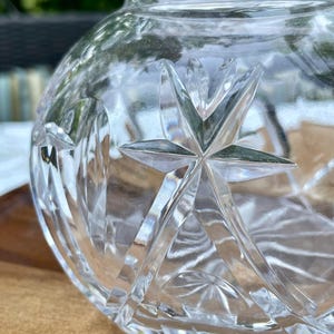 May include: Close-up of a clear, cut-glass decorative vase. The vase features a starburst design with pointed edges and a rounded body. The glass reflects light, creating a sparkling effect. The vase sits on a wooden surface.