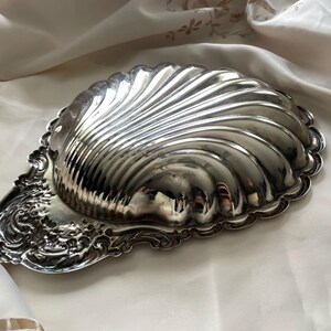 K&ouml;nnte beinhalten: Eine silberfarbene Metallschale in Form einer Jakobsmuschel. Die Schale zeichnet sich durch ein markantes, geripptes Muscheldesign auf dem Hauptk&ouml;rper und ein verziertes, komplexes Schn&ouml;rkelmuster am Griffbereich aus, mit einer polierten, reflektierenden Oberfl&auml;che.