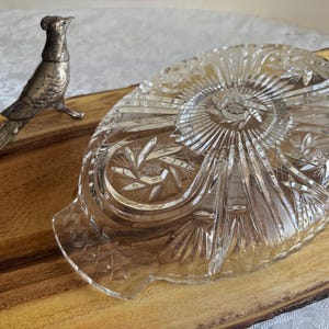 Op de afbeelding: Een heldere, ovale kristallen serveerschaal met ingewikkelde ge&euml;tste patronen, rustend op een houten plank. Een zilverkleurig vogelbeeldje staat links. De schaal heeft een centraal stervormig ontwerp en bloemmotieven, ideaal voor het serveren van hapjes of desserts.