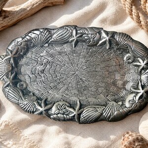 May include: An oval-shaped metallic tray with a dark, antiqued finish. The center features a radiating, textured pattern resembling a spider web or fan, while the raised border is decorated with various seashells, starfish, and sand dollars.