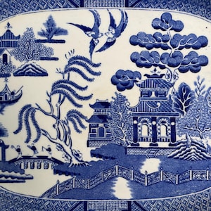 May include: An oval ceramic platter featuring a blue and white Willow pattern. The design depicts a landscape with pagodas, a weeping willow tree, a bridge with figures, a boat on water, and two birds in flight, all framed by a geometric border.