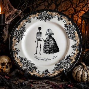 Puede incluir: Un plato redondo de cer&aacute;mica con una ilustraci&oacute;n en blanco y negro de una pareja de esqueletos con ropa de boda. El texto Till Death Do us Part est&aacute; impreso bajo las figuras. El borde tiene un patr&oacute;n floral negro y dorado sobre fondo blanco.