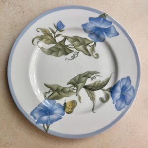May include: A round white ceramic plate featuring a light blue rim and a botanical illustration of blue morning glory flowers, green leaves, and curling vines. A small yellow butterfly is perched on one of the blue flower petals.