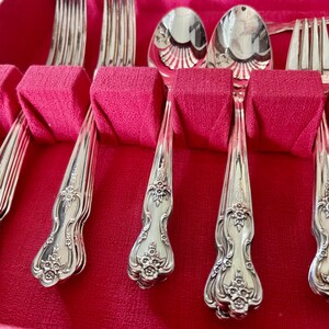May include: A set of silver-toned flatware including forks and spoons arranged in a red storage case. Each piece features an ornate floral embossed pattern on the handle with a polished finish. The utensils are held in place by individual red fabric-covered dividers within the box.