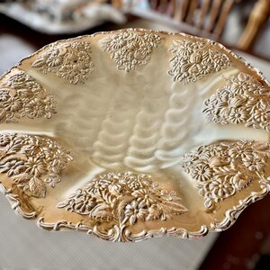 May include: A gold-toned, decorative serving dish with a scalloped edge. The dish features embossed floral designs around the rim and in the center. The interior has a textured, wavy pattern.