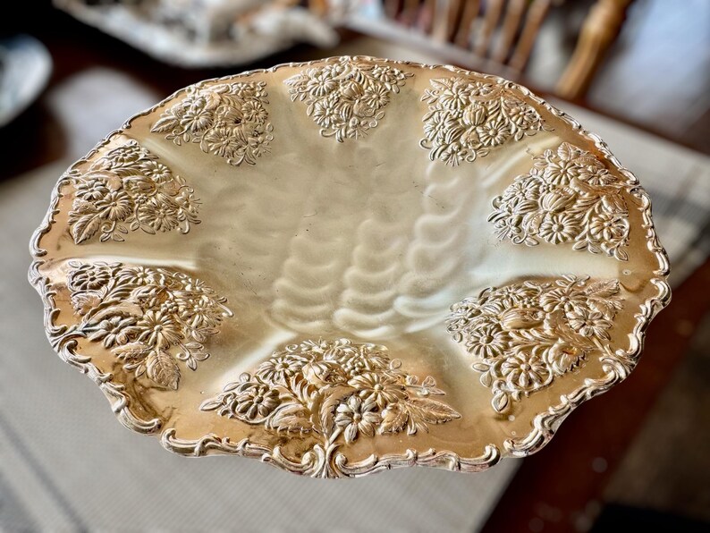 May include: A gold-toned, decorative serving dish with a scalloped edge. The dish features embossed floral designs around the rim and in the center. The interior has a textured, wavy pattern.