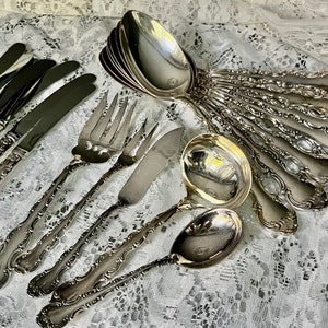 May include: An assortment of antique silverware, including forks, knives, spoons, and a ladle, arranged on a white lace tablecloth. The silverware has ornate, detailed handles and a shiny, reflective surface.