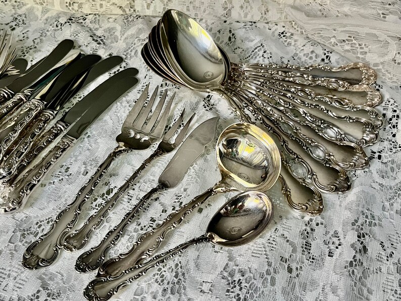 May include: An assortment of antique silverware, including forks, knives, spoons, and a ladle, arranged on a white lace tablecloth. The silverware has ornate, detailed handles and a shiny, reflective surface.