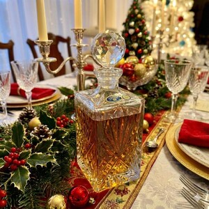 May include: A clear, square-shaped crystal decanter with a faceted stopper, filled with amber liquid. The decanter sits on a table set for a holiday meal, with candles, glassware, and festive decorations.