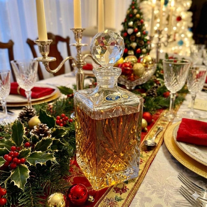 May include: A clear, square-shaped crystal decanter with a faceted stopper, filled with amber liquid. The decanter sits on a table set for a holiday meal, with candles, glassware, and festive decorations.