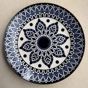 May include: A round ceramic plate featuring a dark blue and white mandala pattern. The design consists of a solid dark blue center surrounded by layers of intricate petal-like shapes, geometric lines, and small black dots on a cream background.