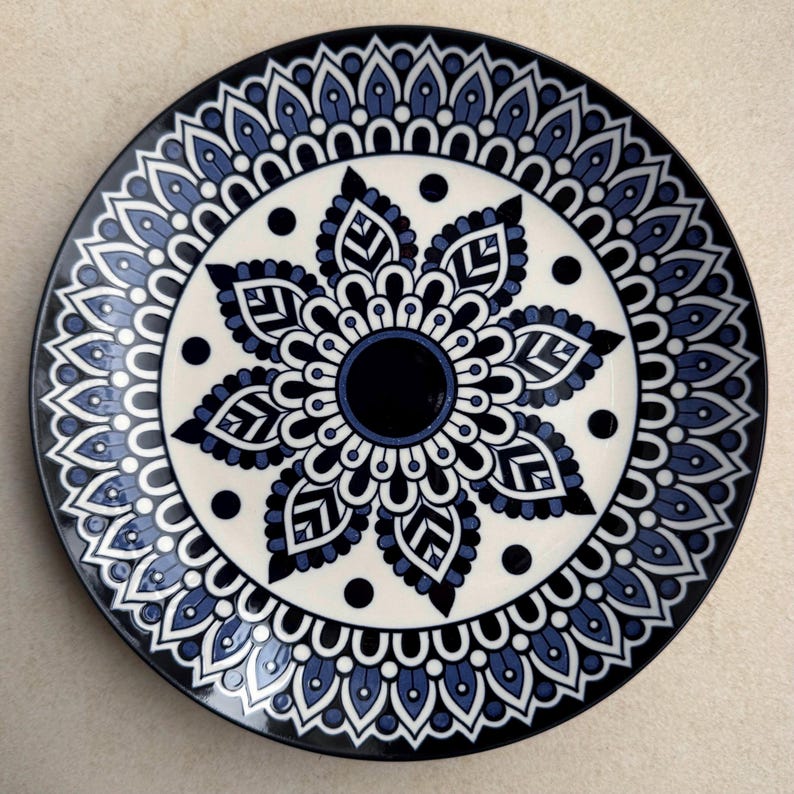 May include: A round ceramic plate featuring a dark blue and white mandala pattern. The design consists of a solid dark blue center surrounded by layers of intricate petal-like shapes, geometric lines, and small black dots on a cream background.