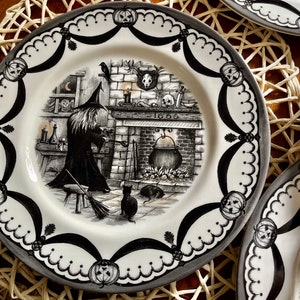 May include: Black and white ceramic dinner plate featuring a witch stirring a cauldron in front of a fireplace. The plate has a decorative border with black and white pumpkins and scalloped edges.