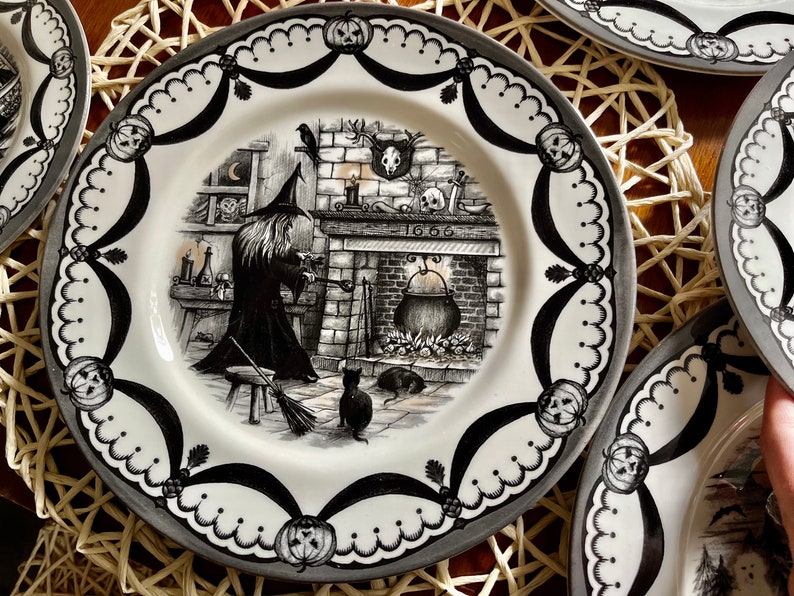 May include: Black and white ceramic dinner plate featuring a witch stirring a cauldron in front of a fireplace. The plate has a decorative border with black and white pumpkins and scalloped edges.