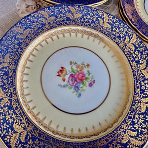 May include: A set of vintage porcelain plates with a floral design. The plates feature a cream-colored center with a floral bouquet and a navy blue border with gold leaf detailing. The plates have a gold rim.