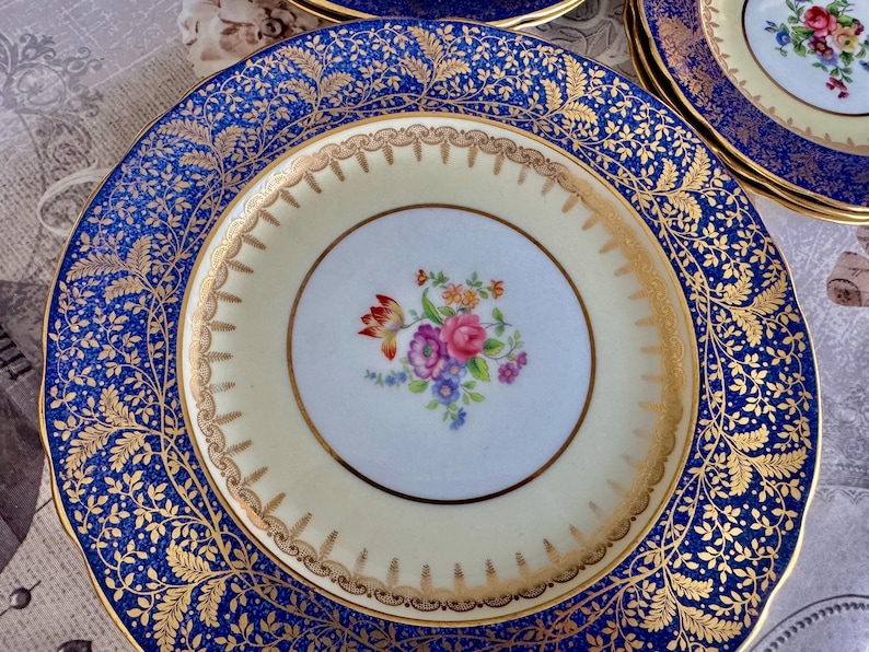May include: A set of vintage porcelain plates with a floral design. The plates feature a cream-colored center with a floral bouquet and a navy blue border with gold leaf detailing. The plates have a gold rim.