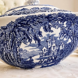 Op de afbeelding: Een witte keramische terrine met een blauw transferware-patroon met een gedetailleerd landschap met een groot kasteel, bomen en figuren. De serveerschaal met deksel heeft een geschulpte rand en een glanzende afwerking in blauw en wit.