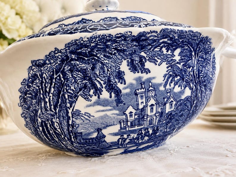 Op de afbeelding: Een witte keramische terrine met een blauw transferware-patroon met een gedetailleerd landschap met een groot kasteel, bomen en figuren. De serveerschaal met deksel heeft een geschulpte rand en een glanzende afwerking in blauw en wit.