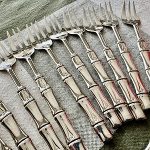May include: A collection of silver forks with bamboo-style handles. The forks are arranged diagonally on a light green and white checkered tablecloth. The tines of the forks are sharp and evenly spaced.