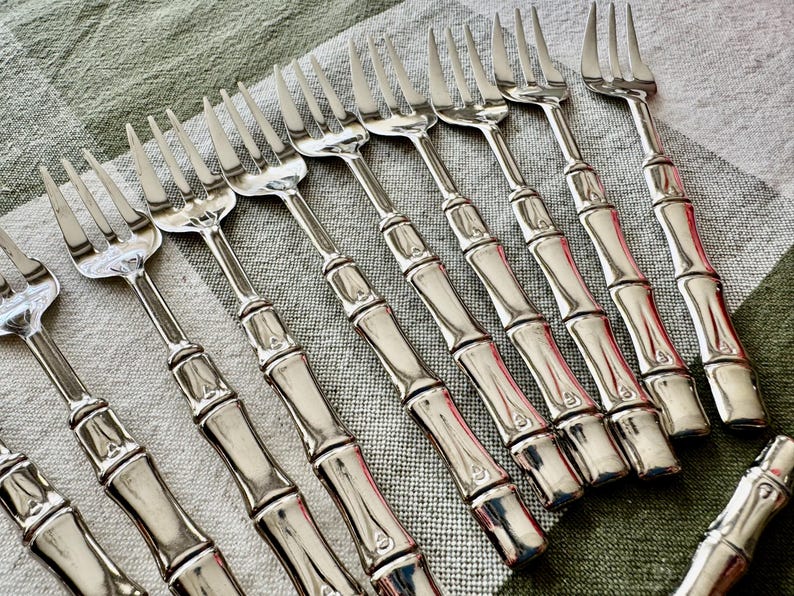 May include: A collection of silver forks with bamboo-style handles. The forks are arranged diagonally on a light green and white checkered tablecloth. The tines of the forks are sharp and evenly spaced.