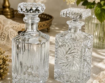May include: Two clear glass decanters with decorative stoppers. One has a round stopper, the other a square. The decanters have textured, geometric patterns. The background includes flowers and a decorative tray.