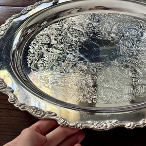 May include: An oval silver-toned serving tray featuring an intricate embossed floral and scrollwork pattern across the center and a decorative scalloped edge with detailed flower motifs along the entire rim of the metallic platter.