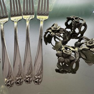 Pu&ograve; includere: Un set di quattro forchette in metallo color argento con manici goffrati a motivi floreali, disposte in fila accanto a tre portatovaglioli in metallo scuro e ossidato con disegni traforati di fiori e foglie con intagli complessi.