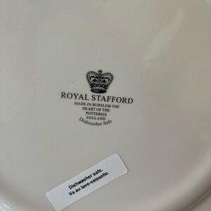 K&ouml;nnte beinhalten: Ein wei&szlig;er Keramikteller mit einem schwarz-wei&szlig;en Logo f&uuml;r Royal Stafford. Das Logo zeigt eine Krone und den Text "Royal Stafford" und "Made in Burslem the Heart of the Potteries England Dishwasher Safe". Ein wei&szlig;er Aufkleber auf dem Teller lautet "Dishwasher safe. Va au lave-vaisselle."