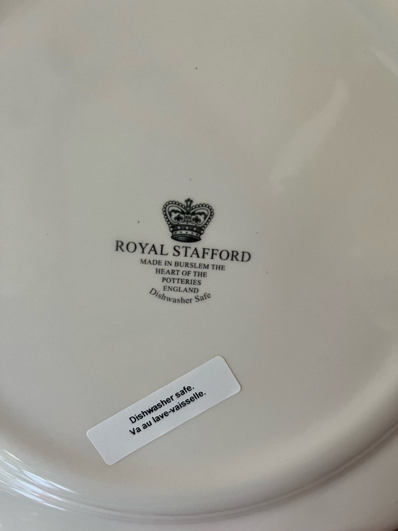 K&ouml;nnte beinhalten: Ein wei&szlig;er Keramikteller mit einem schwarz-wei&szlig;en Logo f&uuml;r Royal Stafford. Das Logo zeigt eine Krone und den Text "Royal Stafford" und "Made in Burslem the Heart of the Potteries England Dishwasher Safe". Ein wei&szlig;er Aufkleber auf dem Teller lautet "Dishwasher safe. Va au lave-vaisselle."