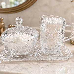 May include: A clear glass vanity set consisting of a lidded bowl, a handled mug, and a rectangular tray, all featuring a faceted starburst pattern. The bowl is filled with white cotton rounds, and the mug contains white cotton swabs.