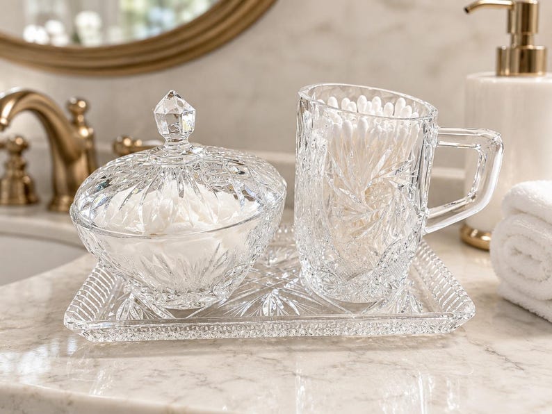 May include: A clear glass vanity set consisting of a lidded bowl, a handled mug, and a rectangular tray, all featuring a faceted starburst pattern. The bowl is filled with white cotton rounds, and the mug contains white cotton swabs.
