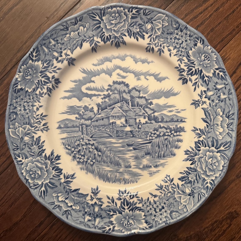 May include: A round ceramic plate with a scalloped edge featuring a blue and white transferware pattern. The center depicts a rural landscape with a stone bridge, a house, and a person, while the border displays a dense floral motif of roses.